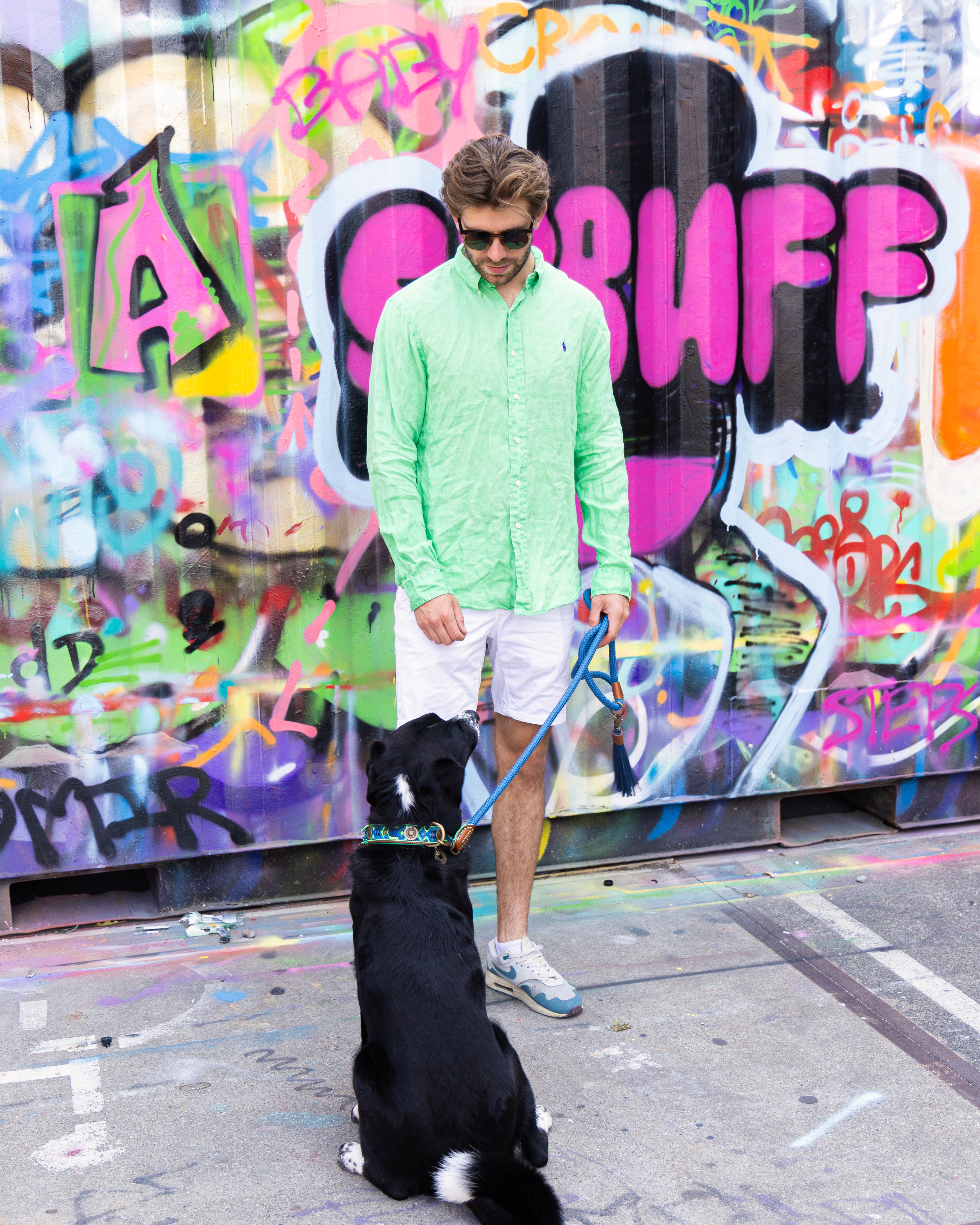 Man in light green shirt and white shorts standing with a dog on a leash in front of a colorful graffiti wall.