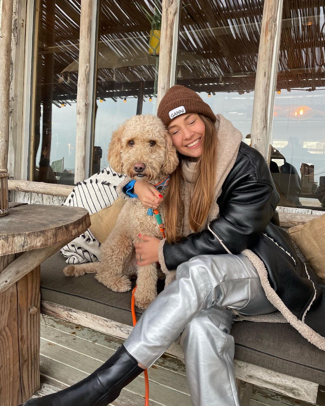 Person sitting on a wooden bench with a dog, wearing a brown beanie and black jacket.